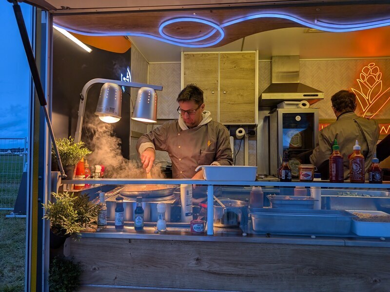 Koch in der Küche eines Foodtrailers beim Kochen, davor Dampf und blaues Neonlicht
