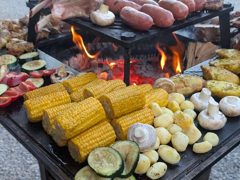 Gemüse, Maiskolben, Champignons und Würstchen auf der Feuerplatte eines Grills