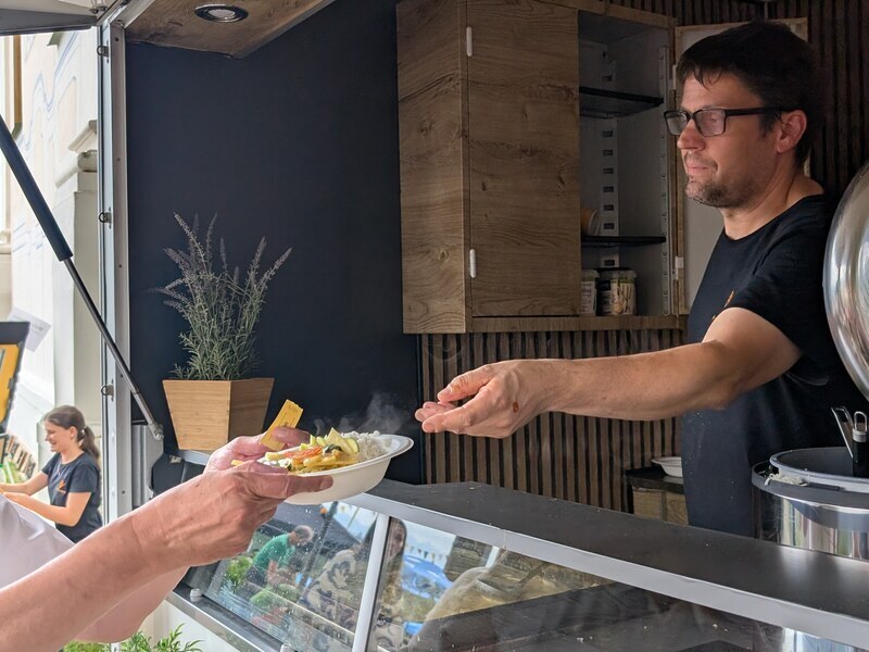 Koch reicht im Foodtrailer einen Teller mit warmem Gericht an einen Gast