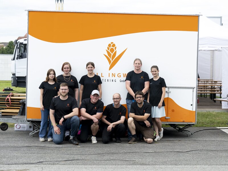 Das Team von Hillingwer Catering posiert vor einem weißen Foodtrailer mit orangem Schriftzug
