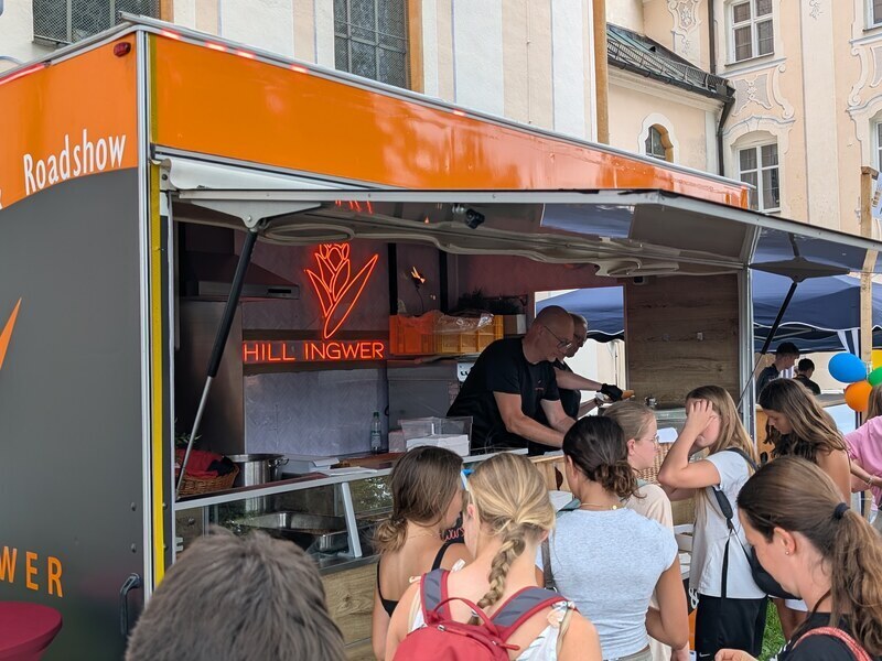 Kinder und Jugendliche stehen am Foodtrailer der Roadshow an, im Hintergrund eine Kirche