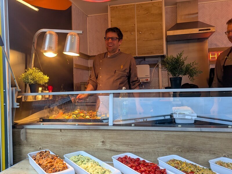 Koch lächelt hinter der Warmhaltetheke des Foodtrailers, vorn Schalen mit Beilagen