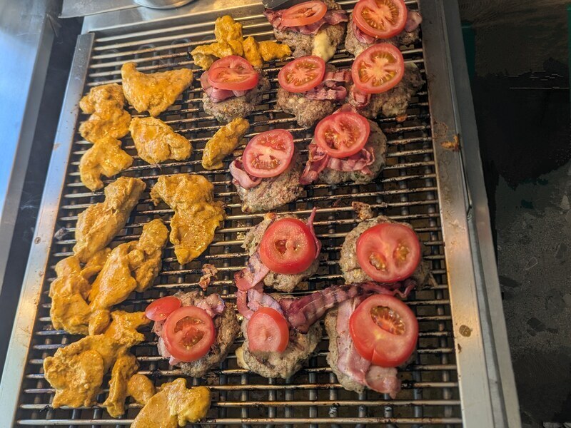 Hähnchenstücke und Burger mit Tomate und Speck auf einem großen Grillrost