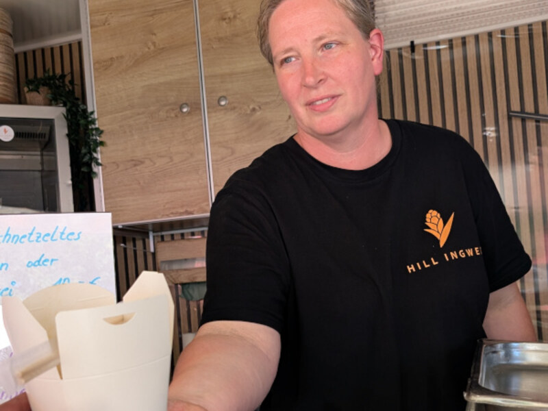 Mitarbeiterin im schwarzen Hill-Ingwer-Shirt blickt aus dem Foodtrailer, davor Take-away-Boxen