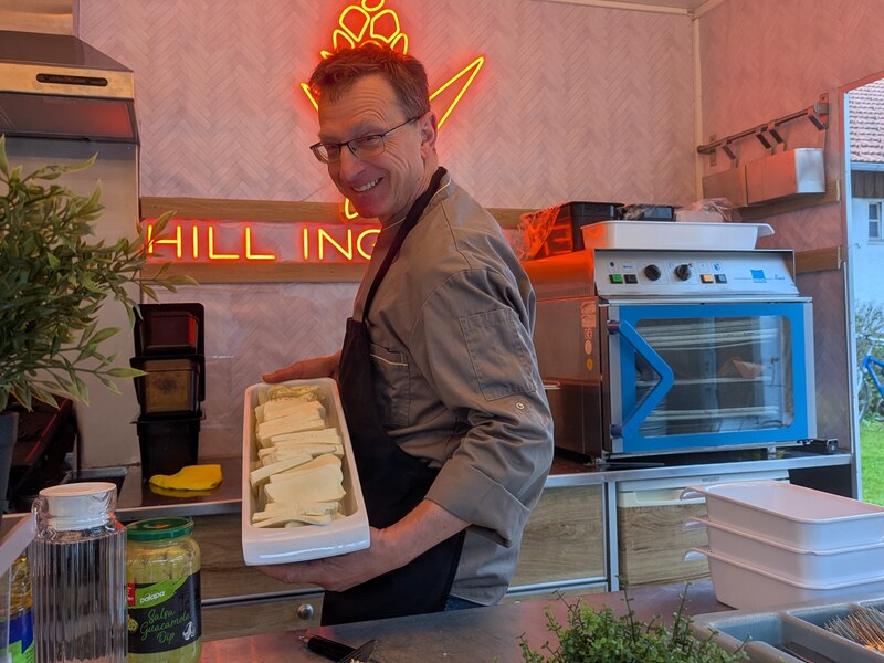 Koch mit Schürze trägt im Foodtrailer ein Blech mit Essen, dahinter Neon-Logo und Ofen