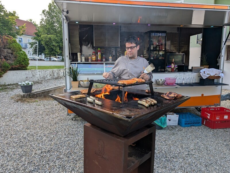 Koch grillt am Feuergrill Würstchen und Fleisch, im Hintergrund der Foodtrailer
