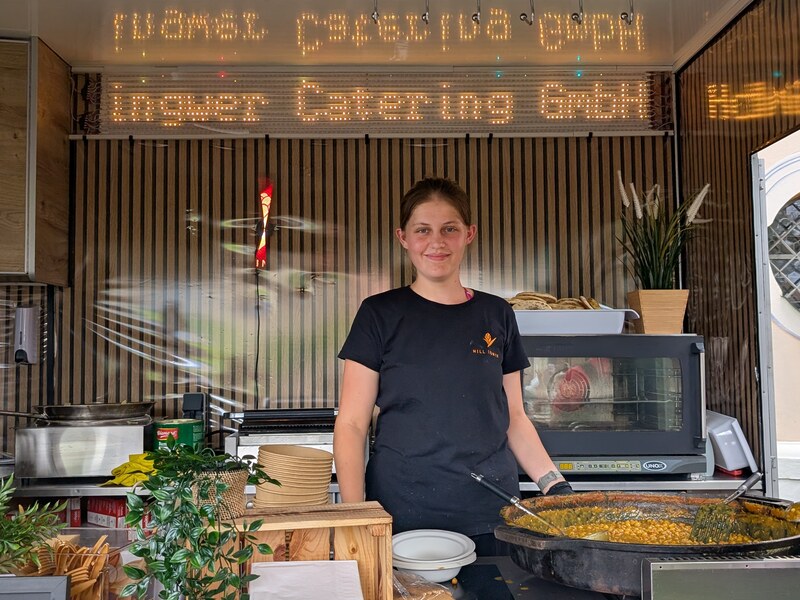 Mitarbeiterin lächelt im Foodtrailer vor einer großen Pfanne und einem Leuchtschriftzug