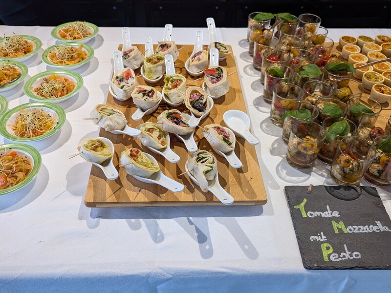 Fingerfood auf Holzbrett und Dessert im Glas auf einem Buffet, daneben ein Schild „Tomate Mozzarella mit Pesto“