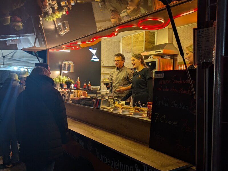Zwei Köche arbeiten am Abend im Foodtrailer, davor Gäste an der Theke mit Tafel