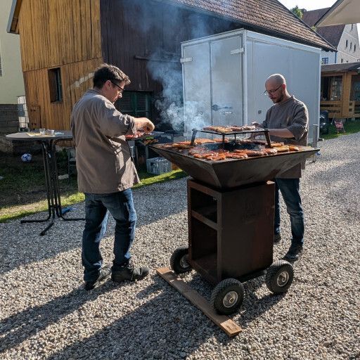 Zwei Köche grillen gemeinsam am fahrbaren Vulcanus-Feuergrill auf einem Kiesplatz