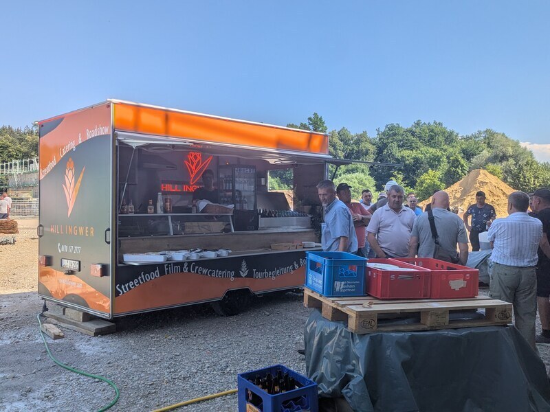 Foodtrailer mit leuchtender Ausgabe auf einem Kiesplatz, davor stehen Gäste