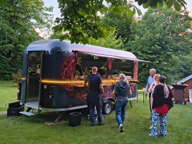 Schwarzer Airbrush-Foodtrailer mit Ausgabeklappe auf einer Wiese, Gäste gehen darauf zu