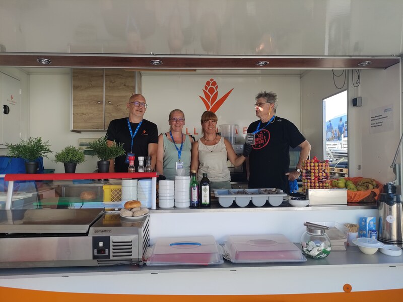 Vier Mitarbeitende lächeln hinter der Theke des Foodtrailers, davor Geschirr und Kühlboxen