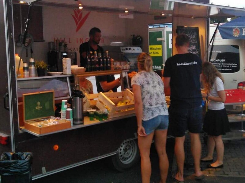 Chefkoch Michael bei der Arbeit im Foodtrailer, davor Gäste an der Theke und eine Tafel mit Tagesangebot