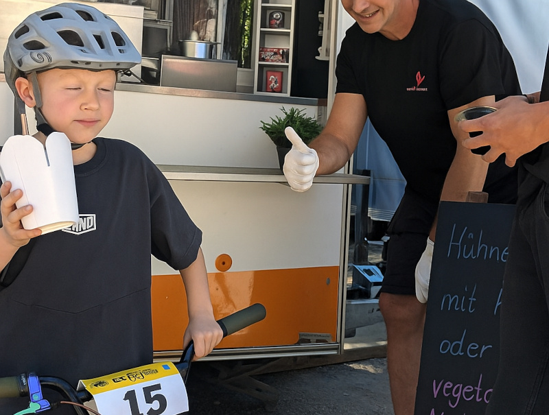 Kind mit Fahrradhelm hält eine Take-away-Box, ein Mitarbeiter zeigt Daumen hoch, daneben eine Tafel